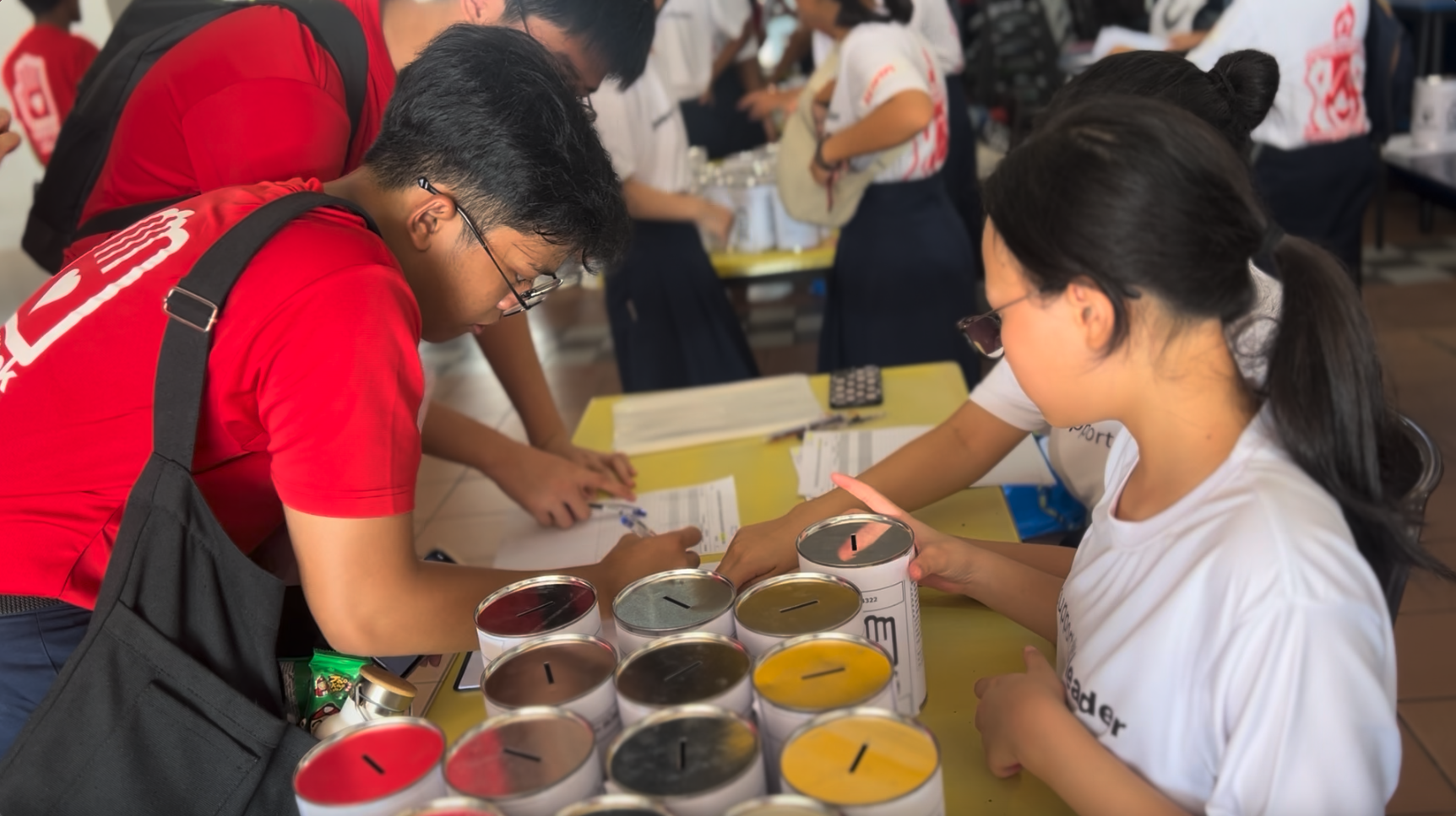 Two people lean over a table filled with colorful donation tins with coin slots at a busy community event.
